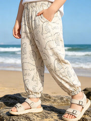 A child wearing beige elastic jogger pants with a black dinosaur and text print, standing on rocks by the beach with the ocean in the background.