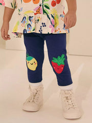 Child wearing navy leggings with fruit designs and a colorful top on a light background