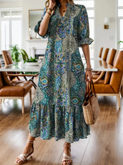 A woman wearing a bohemian-style midi dress with a blue, green, and beige paisley print, holding a woven straw bag in a bright dining room.