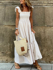 A woman stands against a stone wall wearing a cream and beige gingham maxi dress with ruffled sleeves and a smocked bodice. She is holding a woven straw tote bag and wearing tan sandals.