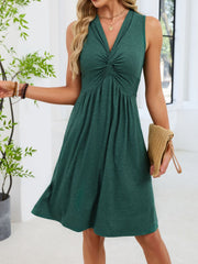 A woman wearing a sleeveless dark green V-neck dress with a gathered twist detail at the bust and a flared skirt, holding a beige woven clutch in a bright indoor setting.